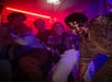 A group of people react with excitement and fear as a person in zombie makeup and costume approaches in a dimly lit, neon-lit setting.