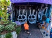 A young boy in an orange shirt aims to throw an object into skull-decorated boards at a carnival game booth with Halloween-themed decorations.
