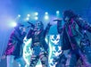 Three musicians in leather jackets perform on stage with microphones, colorful lights, and a digital screen with graphic designs in the background.