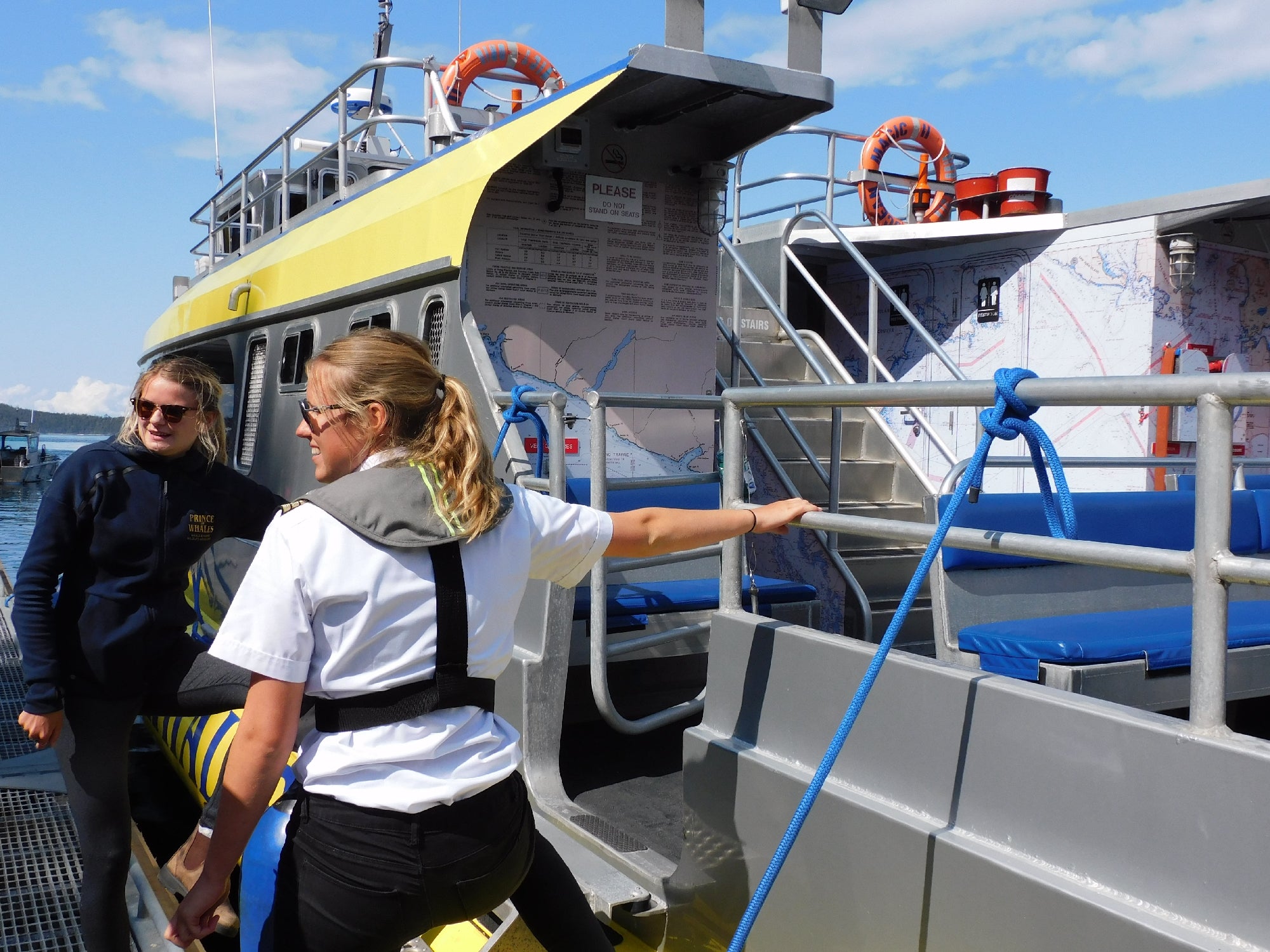 Two people stand on a dock next to a yellow and gray boat, one person holds onto a rope attached to the vessel.