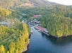 Aerial view of a small waterfront town surrounded by dense forest, with red buildings and boats docked in a marina along a narrow inlet.
