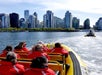 People in red jackets ride a yellow boat on a city waterfront, with tall buildings and another boat visible ahead under a clear sky.
