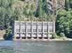 A concrete hydroelectric power station with five large arched windows stands by a forested, rocky hillside at the water's edge.