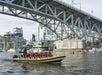 A yellow inflatable tour boat with passengers in red life jackets travels under a large steel bridge in an urban waterfront area.
