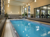 Indoor swimming pool with tiled floor, surrounded by large windows. Pool is illuminated by overhead lights, with lounge chairs and plants along the sides. Gym equipment visible in adjacent room.