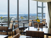 A bright cafe with wooden tables and chairs, yellow flowers on each table, and large windows offering a scenic view of a city and distant hills.