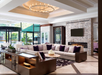 Modern living room with a large sectional sofa, accent pillows, and a central coffee table. Features a fireplace, wall-mounted TV, and chandelier. Large windows provide natural light.