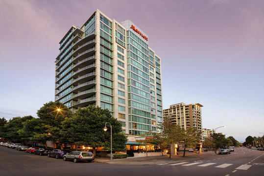 Street view of a tall hotel building with a lit sign and surrounding trees, cars parked along the street, and a nearby crosswalk.