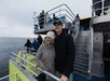 A couple stands on the deck of a boat, overlooking the ocean. Other passengers are visible in the background.