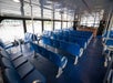 Interior of a ferry with rows of unoccupied blue seats facing forward and large windows on the sides. A counter is visible at the front, and the floor is covered with dark carpet.