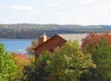 A wooden house is surrounded by lush trees with autumn foliage, overlooking a calm lake and distant hills under a cloudy sky.