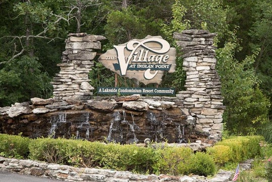 Stone sign for "The Village at Indian Point" with a waterfall feature, surrounded by trees and shrubs.
