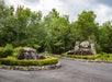 Entrance with a circular driveway, featuring two stone waterfalls and a sign reading "The Villages" surrounded by lush greenery. An American flag flies on a pole to the left.