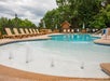 Outdoor swimming pool with a shallow area featuring fountains, surrounded by lounge chairs and umbrellas, set against a backdrop of trees and a cloudy sky.