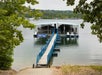 A wooden walkway leads to a covered dock with boats on a calm lake, surrounded by trees.