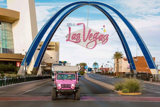 A pink Jeep drives under a large “Las Vegas” sign with blue arches on a sunny day in an urban street setting.