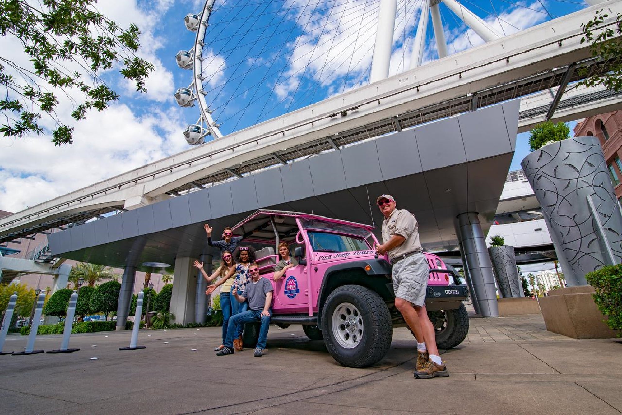 A group of people pose with a pink jeep parked under a large Ferris wheel structure on a sunny day.
