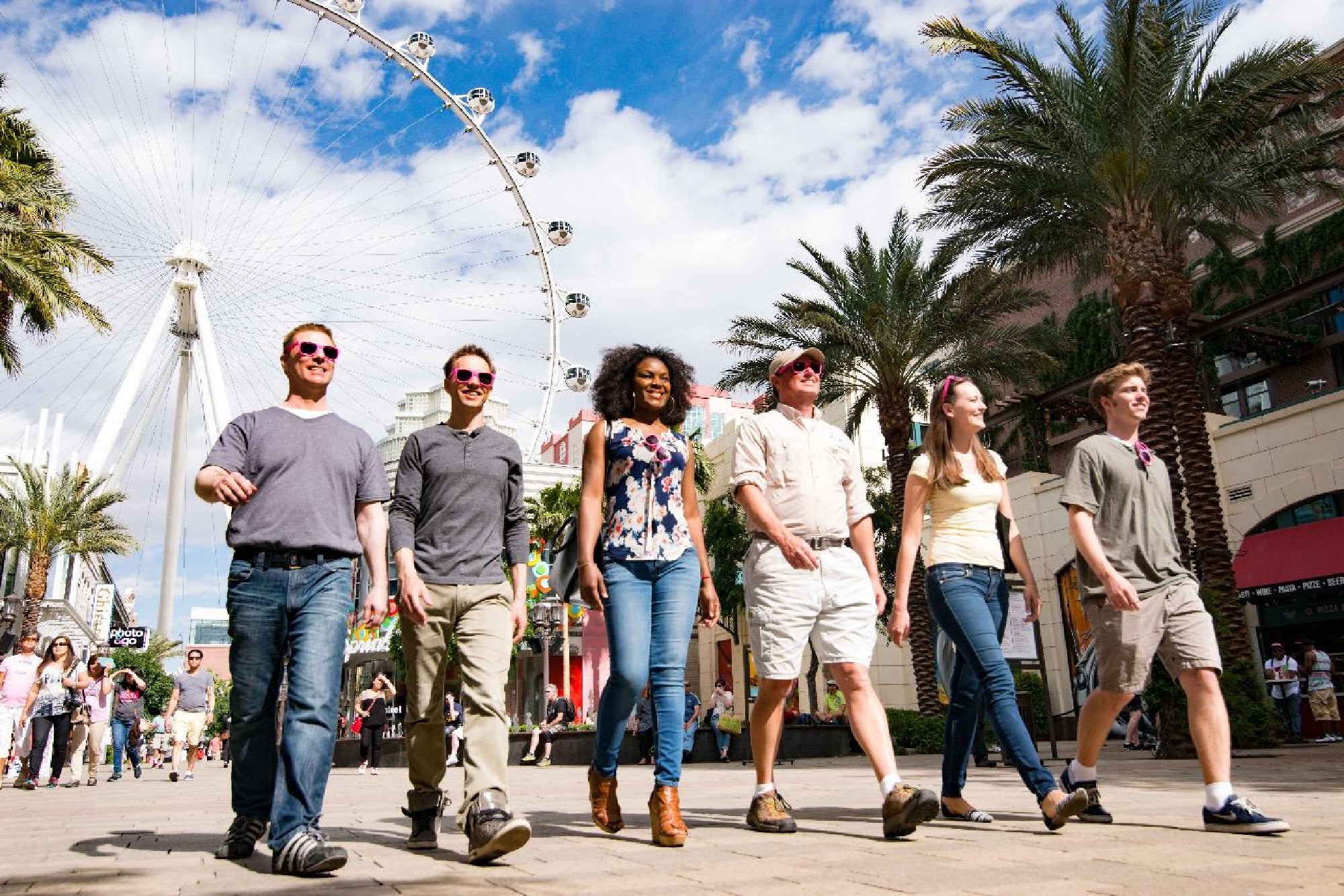 Six people walk together outdoors with palm trees and a large Ferris wheel in the background under a partly cloudy sky.