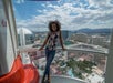 A woman stands inside a glass observation pod with a view of Las Vegas hotels and the city skyline in the background on a sunny day.