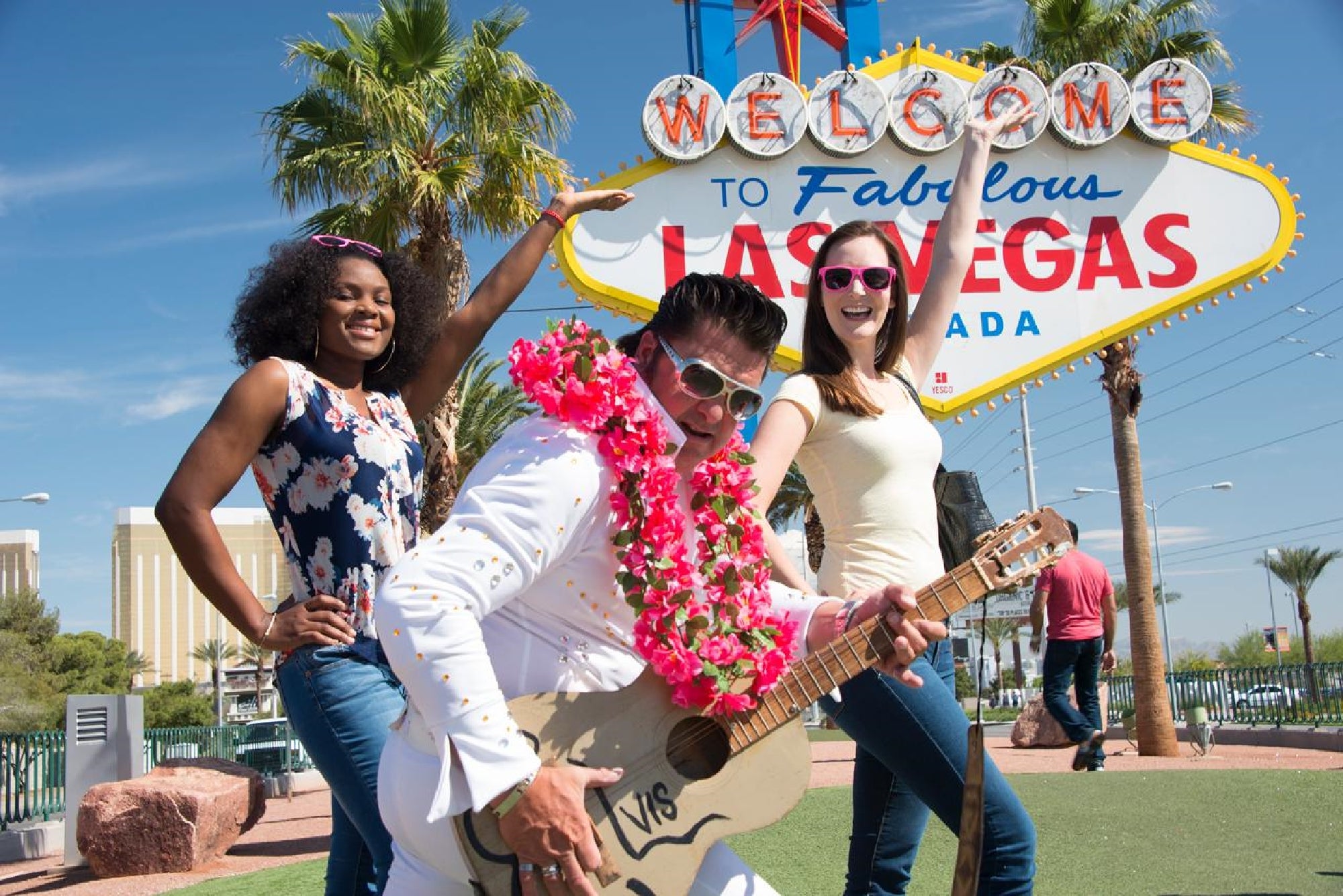Three people pose in front of the "Welcome to Fabulous Las Vegas" sign, including a man dressed as Elvis with a guitar and two women smiling beside him.