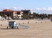 A sandy beach with a lifeguard tower and several buildings in the background. Few people are walking along the shore. Palm trees and trash cans are visible.