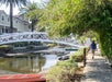 Person walking on a path beside a canal with a white bridge and greenery. A red kayak is in the water under the bridge. Palm trees and a house are visible in the background.