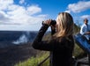A woman uses binoculars to observe a volcanic crater emitting smoke, with another person standing near an information sign in the background.