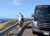 Three people stand by a guardrail at a coastal viewpoint, with one person pointing toward the ocean; a black Mercedes van is parked nearby.