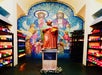 A statue of a saint holding flowers stands before a colorful mosaic of religious figures, surrounded by rows of lit votive candles on both sides.