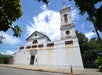 A white church with a tall bell tower stands on a quiet street under a partly cloudy sky, with trees and power lines nearby.