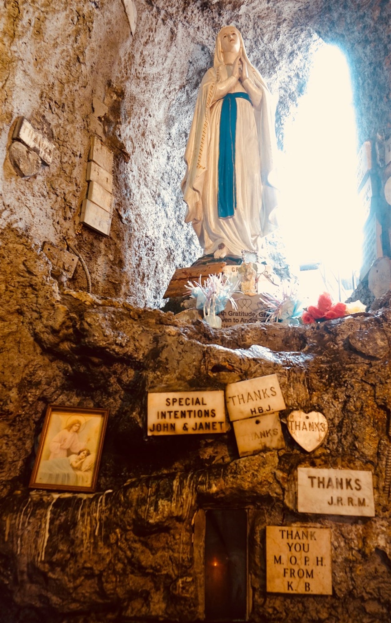Statue of the Virgin Mary in a stone alcove, surrounded by plaques and signs expressing thanks and special intentions, with a framed photo and flowers nearby.