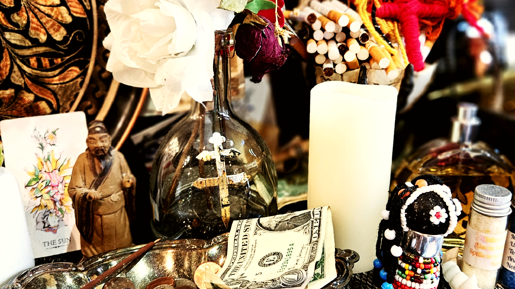 A tabletop displays a $1 bill, a candle, a glass bottle with crosses, dried flowers, a figurine, a tarot card, beads, and various small objects.