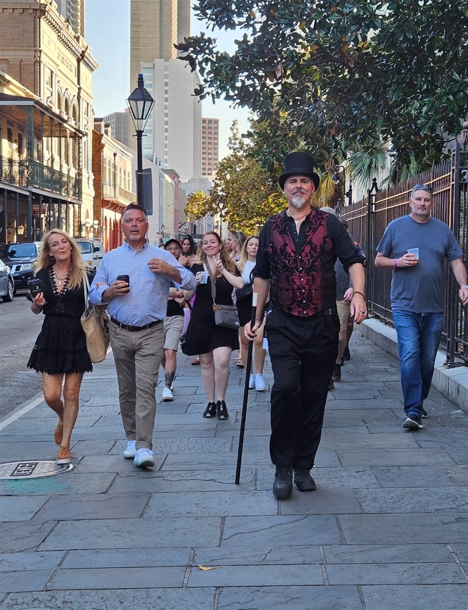 A group of people walk down a city sidewalk; a man in front wears a red vest, black hat, and carries a cane. Buildings and trees line the street.