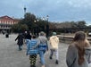 A group of people walk along a paved path near a black iron fence and historic buildings under a cloudy sky.