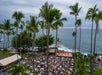 Aerial view of an outdoor event near the ocean, with tables set up and a performance stage among tall palm trees.