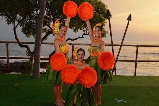 Three performers in traditional costumes hold bright orange pom-poms against a backdrop of the ocean and sunset.