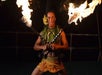 A performer in traditional Polynesian attire holds two flaming torches crossed in front of him during a fire dance at night.