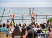Two performers in traditional attire hold torches on stage by the ocean, entertaining a seated audience at an outdoor event.