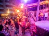Performers in traditional clothing, holding torches, walk past an audience at an outdoor night event with live music and stage lighting.