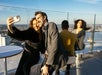 A group of people on a rooftop terrace, two taking a selfie while others sit and enjoy the view.