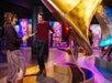 Two people dance joyfully near a large, golden gramophone in a colorful, art-filled room. Other visitors are seen in the background.