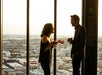 Two people converse near tall windows overlooking a cityscape, each holding a drink.