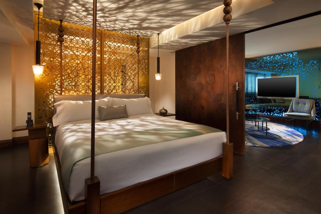 A modern hotel room with a large bed, decorative lattice wall, pendant lights, and a separate seating area with a TV and chair.