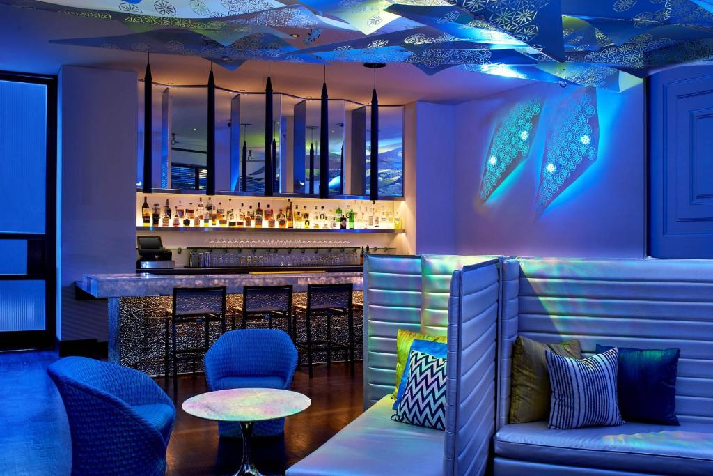Modern lounge bar with blue ambient lighting, geometric ceiling design, and a white marble bar. Features seating with round tables, cushioned booths, and a display of illuminated bottles.
