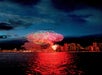 Fireworks light up the evening sky above city buildings, reflecting colorful lights on the water below.