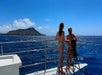 Two people in swimsuits stand on a boat deck overlooking the ocean, with a mountainous island and blue sky in the background.
