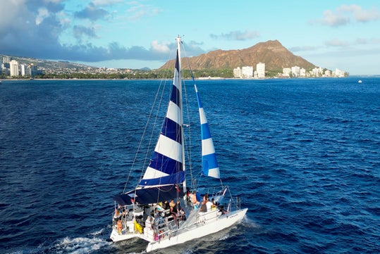 A white sailboat with blue and white striped sails sails on the ocean near a city shoreline, with a mountain and buildings visible in the background under a blue sky.