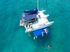 Aerial view of a catamaran anchored in clear blue water with people swimming and relaxing on floating mats around the boat.
