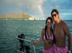 A smiling couple in pink swimwear stands on a boat with the ocean, a city skyline, a mountain, and a rainbow in the background.