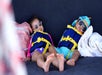 Two young children wearing life jackets, swimsuits, and sunglasses are lying down and resting next to a towel and a bag.